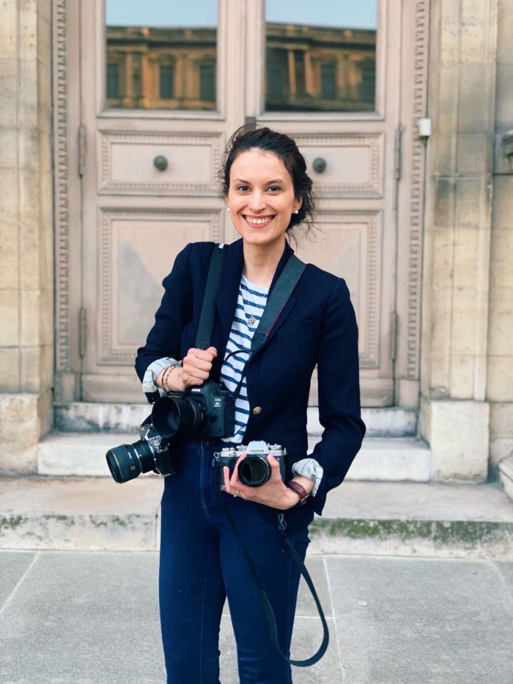 A Caucasian woman wearing a blue blazer and blue jeans, with her brown hair up in a bun, holding 3 cameras