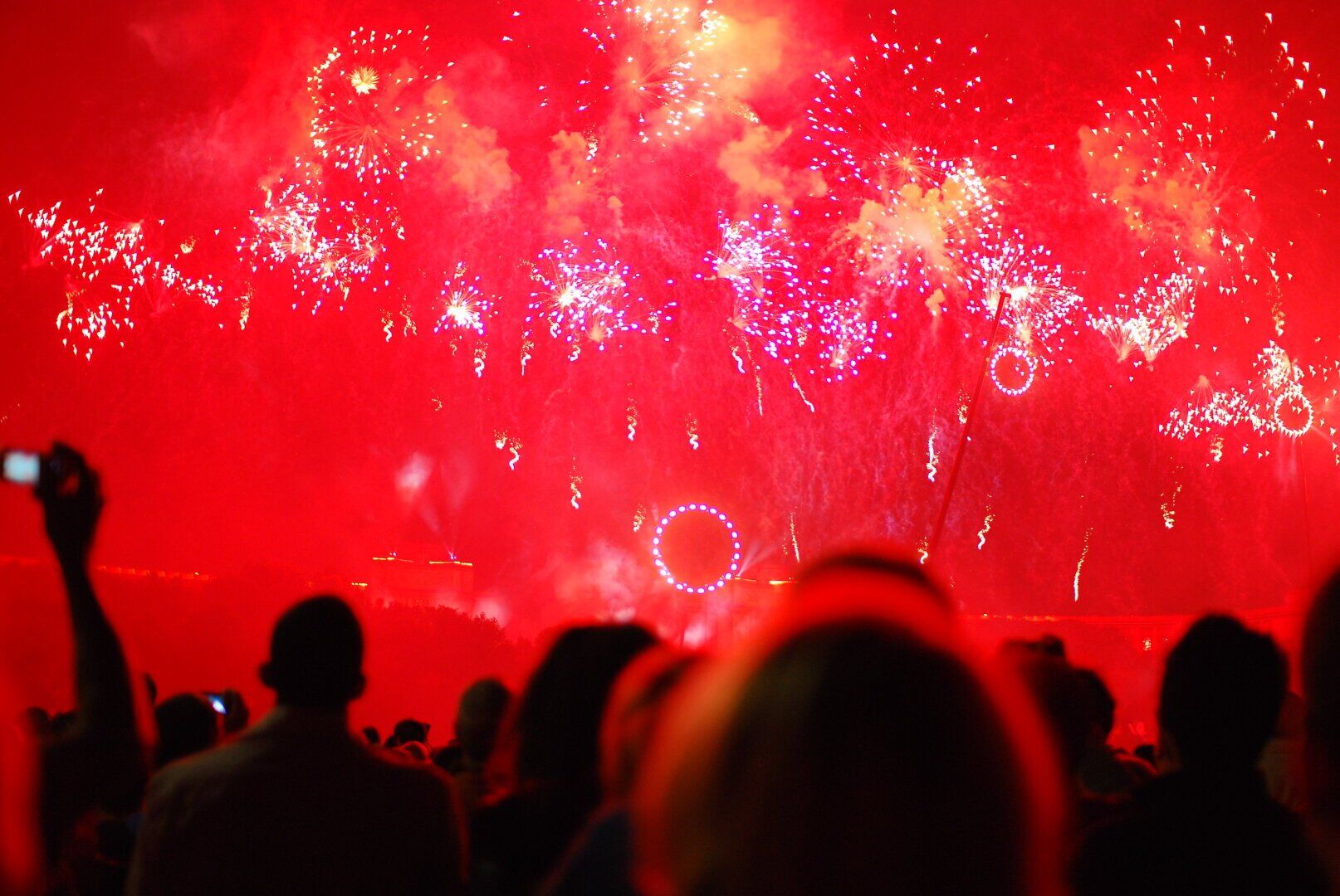 a photo showing a bright red sky with sparkles of fireworks and below you can see the silhouette of people looking up and a cell phone capturing the scene