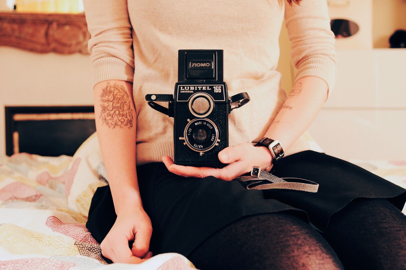 a photo of a woman cradling in her lap a Lomo Lubitel 166. You can't see the woman's face, only her upper body. She's sitting on a bed, with a fireplace behind her. On her arm there's a temporary tattoo with the message 