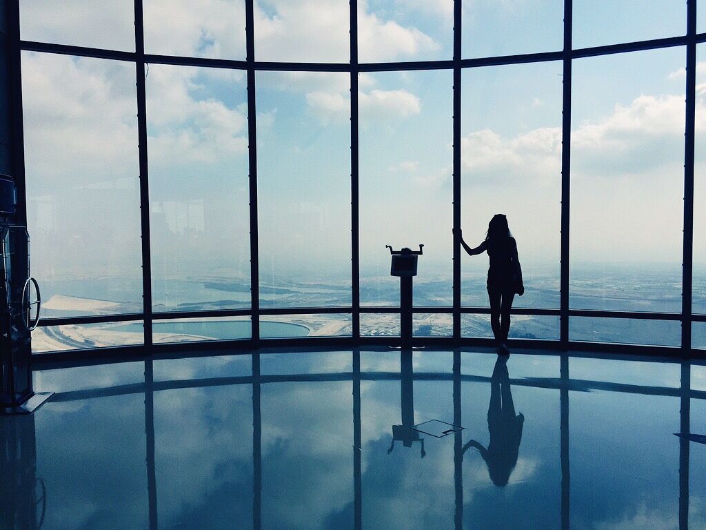 the silhouette of a woman (me) seen from the back as the looks out - from the observation deck of the Burj Khalifa in Dubai, one of the world's tallest buildings. The photo is bathed in blue tones and you can see tall large window panes