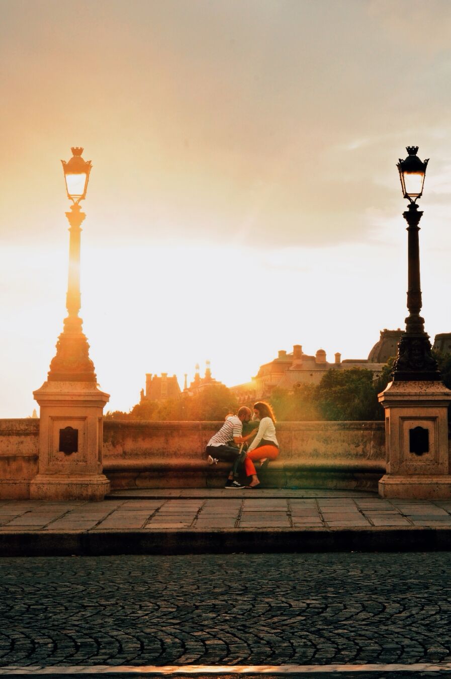 a photo of two lovers sitting across each other on the bridge Pont Neuf at magic hour, with golden orange hues in the sky. The man is gently placing his hand on the woman's chest, the woman is looking down